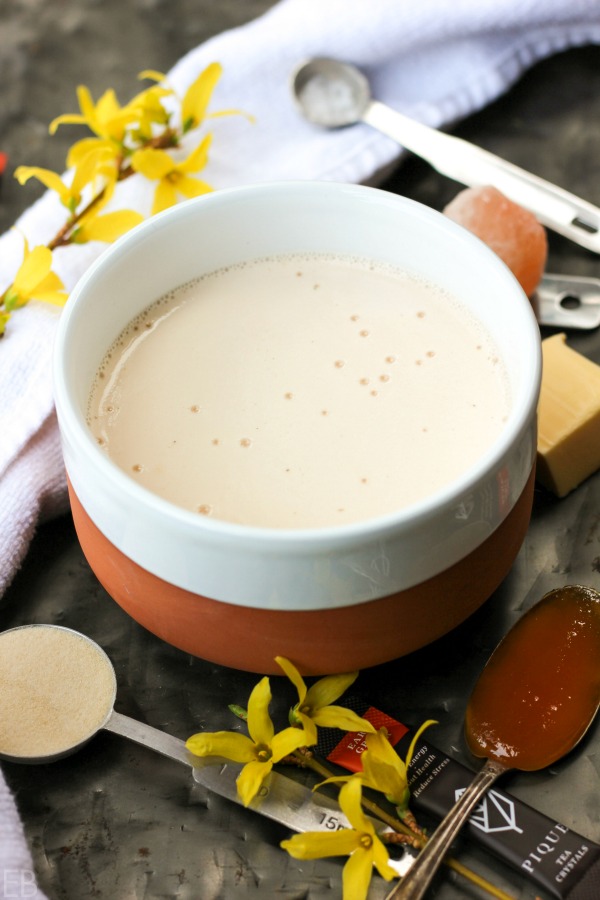 a bowl mug of creamy butter tea surrounded by honey, flowers, tea, salt and gelatin