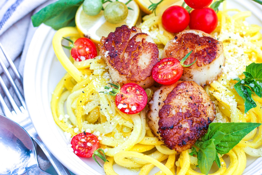 side image of scallop scampi with zucchini noodles, garnished with parmesan, tomatoes and basil