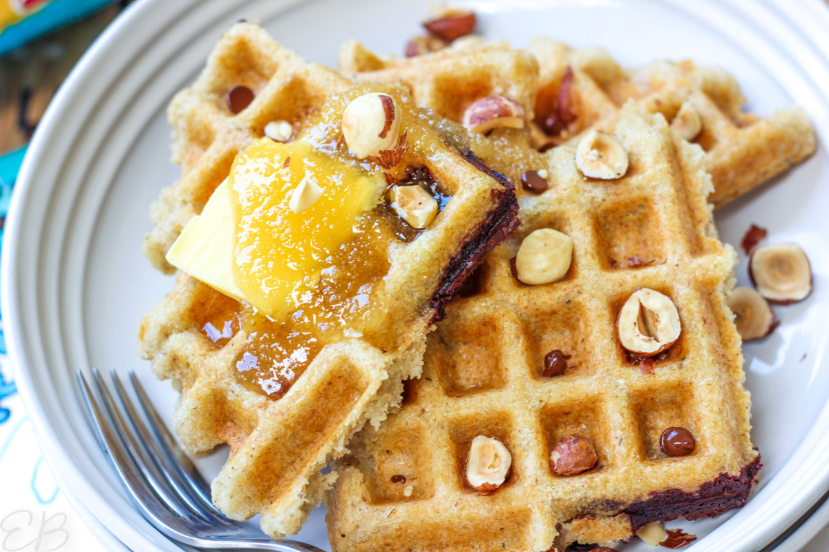 several bean and oat waffles piled on a plate with butter, honey, hazelnuts and chocolate chips on top