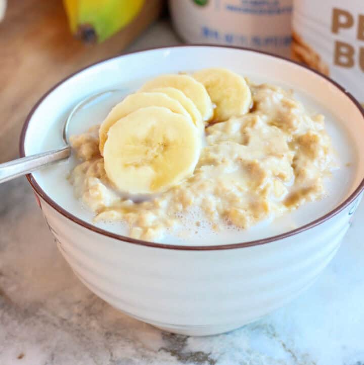 side angle view of gluten free vegan peanut butter banana oatmeal with spoon, banana slices, milk and maple syrup