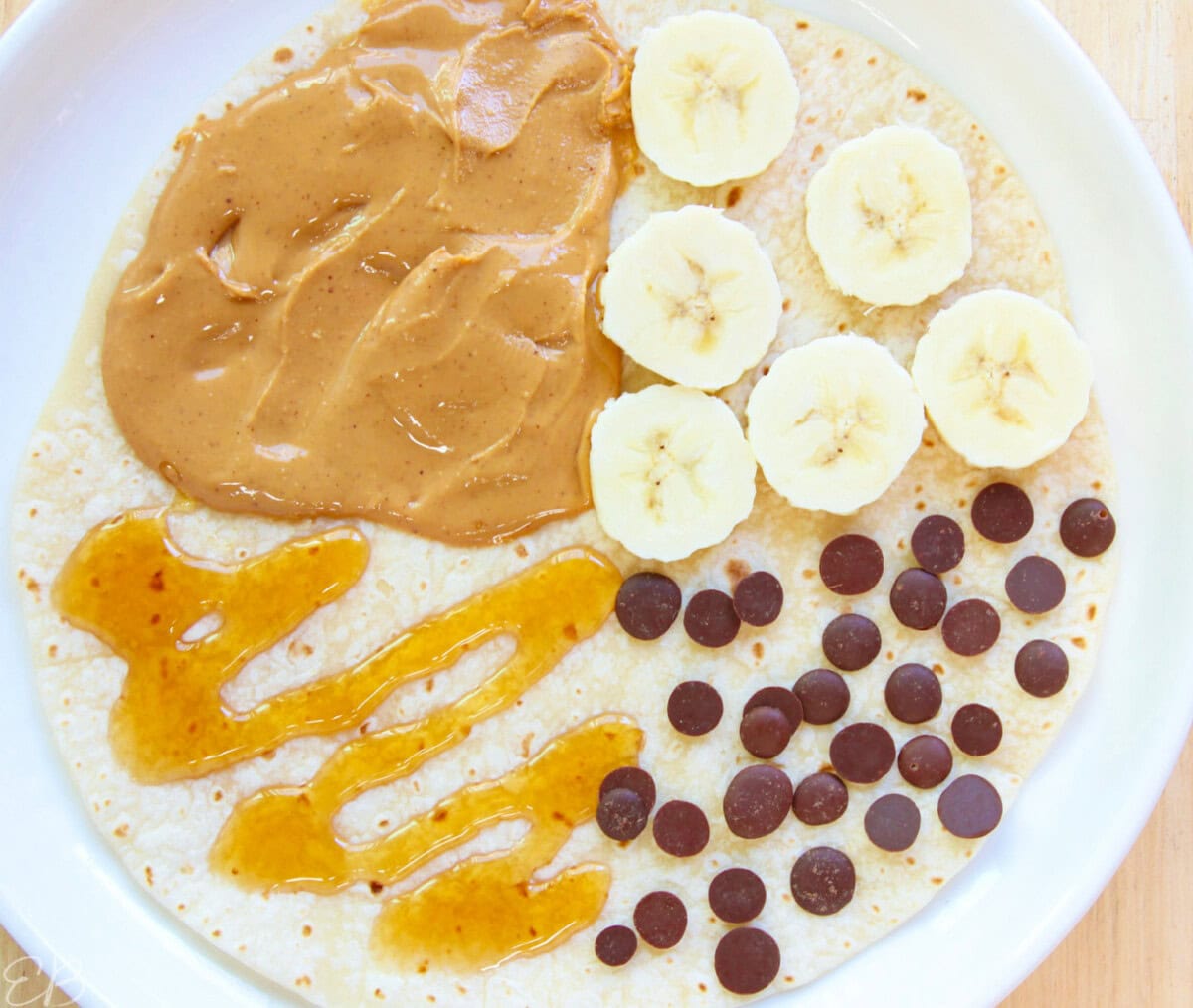 Overhead view of peanut butter (or almond butter), banana, honey and chocolate chips wrap for kids' lunches, before folding over