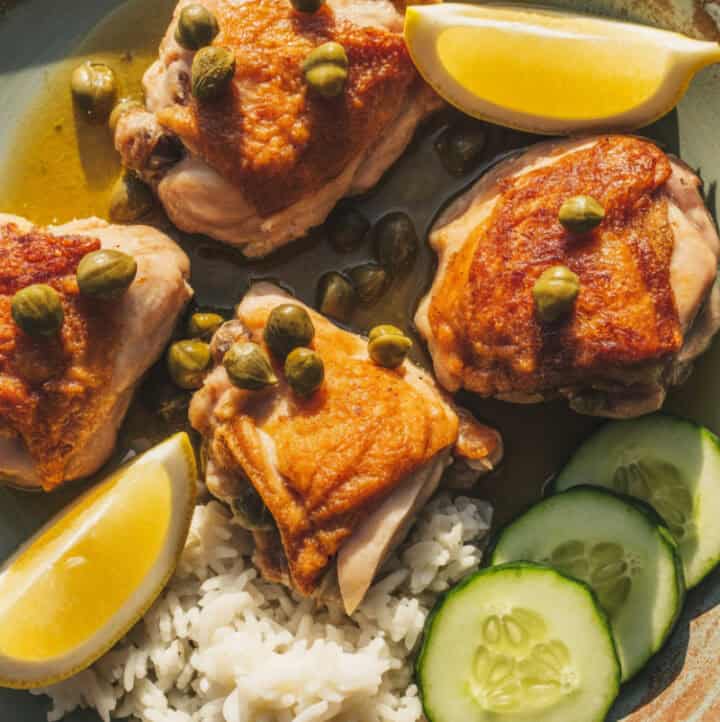 overhead view of baked chicken piccata, garnished with lemon slices and capers, with rice