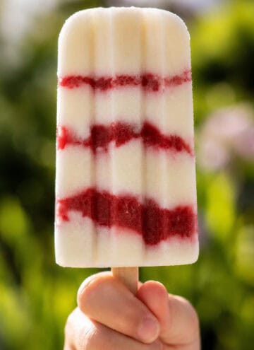 a child's hand holding up one vegan cheesecake popsicle with strawberry layers, outside in summer