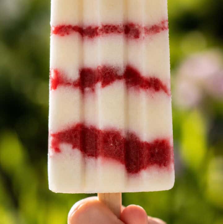 a child's hand holding up one vegan cheesecake popsicle with strawberry layers, outside in summer